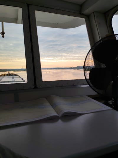 The image shows a view from inside what appears to be a ship's bridge, looking out over a body of water with land visible in the distance. A navigational chart is open on a table in the foreground, and a fan is visible to the right. The sky suggests it is either sunrise or sunset. The location is possibly near Нифантово, Russia.