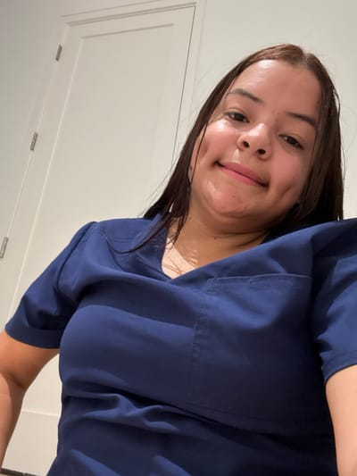 In this close-up, rotated selfie, a young woman with dark hair smiles, her left dimple accentuated. She is wearing a dark blue scrub top, a common garment for healthcare professionals. The background consists of a plain white wall, suggesting an indoor setting such as a home or a medical facility. The lighting appears to be artificial, casting soft shadows and highlighting her skin. The overall mood conveyed is casual and friendly. There is no visible text in the image. The location context provided is Dallas, United States of America.