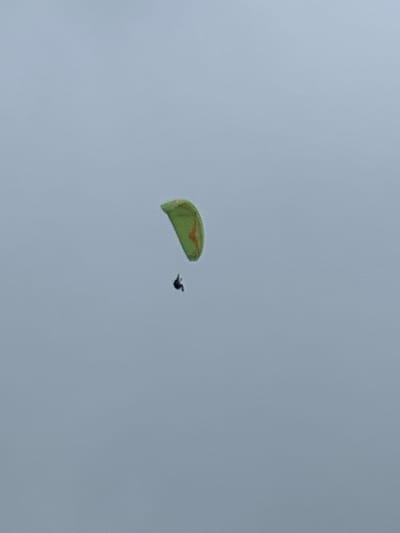 An image captured in the city of Puncak, Indonesia, depicts a single paraglider suspended against a uniformly gray, overcast sky. The paraglider's canopy is light green with faint orange or reddish markings. A solitary, dark human silhouette, the paraglider pilot, is visible directly beneath and attached to the canopy. The individual is actively engaged in paragliding, airborne at a significant altitude, with no ground features or other objects visible in the frame.