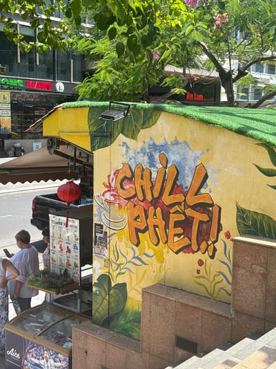 This image captures a street scene in Nha Trang Ward, Vietnam, likely on a sunny day. The foreground features a food stall painted with vibrant yellow and green colors, adorned with a mural that says "CHILL PHÊT!" in large, bold orange and yellow lettering. The mural also includes drawings of leaves, flowers, and a steaming cup. To the left of the stall, there is a display of fresh fruits, including pineapples and other produce, and a glass-fronted freezer with an advertisement for "Aice" featuring a soccer player.
In the background, a black pickup truck is parked, and a red lantern hangs from the stall's awning. A menu board for "Smoothie Fruit Juices" is visible, listing various flavors. Two women are present near the stall; one, in the foreground, is wearing a white top and patterned pants, and is looking at her phone. The other woman, further back, is wearing a patterned top and is partially obscured. Behind the stall, a building with glass windows and signage reading "An Lam RESTAURANT" is visible, suggesting a commercial area. Trees with lush green leaves and some pink blossoms provide shade. The overall impression is a lively urban setting with a focus on a refreshing beverage stand.