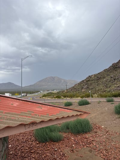 The image depicts a roadside landscape under a cloudy sky, with a view of mountains in the distance. In the foreground, a section of a weathered, corrugated red metal roof is visible, along with piles of reddish-brown gravel and patches of green, bushy vegetation. A lamppost stands to the left of the frame. Beyond the immediate foreground, a highway curves through the scene, with several trucks and cars visible on the road. The terrain is arid, characterized by sparse vegetation and rocky hillsides. The overall impression is one of a remote, possibly desert, environment. The location is stated to be the city of Etholen, United States.