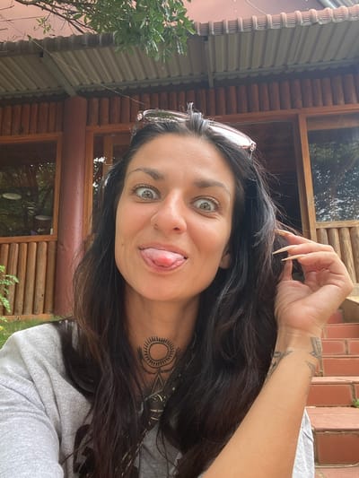 A medium shot captures a woman with dark hair and a playful expression, sticking her tongue out at the camera. She is wearing a grey t-shirt and sunglasses perched on her head. A tattoo is visible on her neck. The background features a rustic building with wooden beams and a corrugated metal roof, suggesting an outdoor setting. There are also some steps made of red brick visible in the background. The environment appears to be natural, with greenery visible around the building. The location is not specified as Thôn 2, Vietnam, as this information is not derivable from the image.