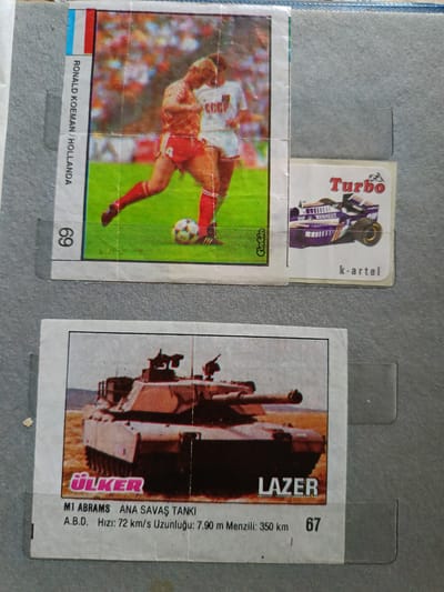 This media file contains images of three collectible cards. The top left card features a soccer player, Ronald Koeman, in action on a field, identified with the text "RONALD KOEMAN / HOLLANDA". The top right card displays a purple Formula 1 race car with the word "Turbo" prominently displayed, along with the text "k-artel". The bottom card depicts a modern tank, identified as the "M1 ABRAMS ANA SAVAŞ TANKI" with details about its speed and length. The text "LAZER" is also visible on this card. The cards appear to be part of a collection, possibly sports or military-themed. The background suggests they are placed on a textured surface, possibly a binder or album page. The lighting is consistent across the images, indicating they were photographed in a well-lit indoor environment. There are no specific time of day or weather cues, and the location context is not discernible from the images themselves. The overall presentation is that of a display of collectible items.