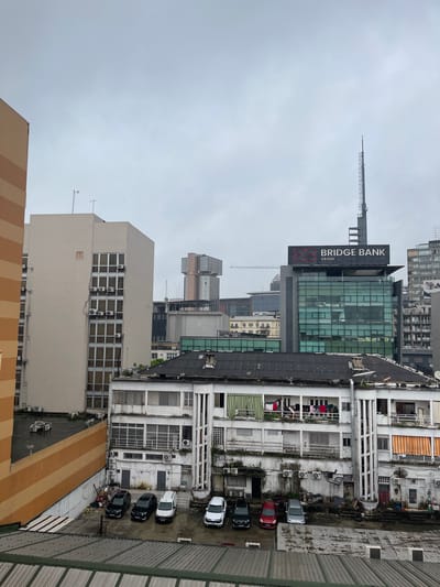 This is an aerial, medium shot of an overcast urban environment in Abidjan, Ivory Coast. The scene is dominated by various buildings, ranging in architectural style and height, suggesting a dense city landscape.

In the foreground, a low-rise white building with multiple stories features a row of balconies on the upper floors, some adorned with colorful laundry hanging out to dry. Below these balconies are numerous windows and doors. The lower levels of this building appear to be garages or storage areas, with several cars parked outside, including black sedans, a white van, and a silver car. Piles of debris are visible near the lower entrances of the building.

Adjacent to this building, on the left, is a taller, light-tan colored building with several windows and what appear to be air conditioning units mounted on its exterior. To the far left, a portion of an orange and tan striped building is visible.

Further in the background, a modern glass-fronted building with a prominent "BRIDGE BANK GROUP" sign stands out. Above this building, a tall, slender communication tower rises into the sky. Other buildings of varying designs and heights are scattered throughout the middle and far background, including a distinctive tiered building in pale pink and brown.

The sky is overcast and appears to be a uniform light gray, contributing to a subdued lighting condition across the scene. The presence of multiple antennas and satellite dishes on the rooftops indicates a populated urban area. The overall impression is one of a functional, lived-in city with both older and newer structures.