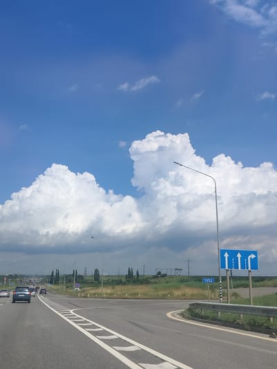 The image depicts a highway scene on a bright, cloudy day, likely in Labinsk, Russia, given the road signage with the number "1352". In the foreground, a paved road curves to the left. White lane markings are visible, including solid and dashed lines. A metal guardrail runs along the right side of the road. A blue traffic sign with white arrows indicates that the left lane goes straight, the middle lane goes straight, and the right lane turns right. A streetlamp stands next to the sign. In the background, there are fields of grass and scattered trees, with power lines and pylons visible. Several cars are driving on the highway, with some appearing to be in a line. The sky is a vibrant blue with large, fluffy white clouds.