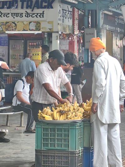 The image captures a street scene in Khandwa, India, likely a market or near a food establishment, judging by the signage and presence of food items. In the foreground, a vendor, wearing a white shirt and a black cap, is actively arranging a large display of bananas on a metal plate atop two stacked green and gray plastic crates. To the right of the vendor, an older man, distinguished by his bright orange turban and white attire, stands observing the bananas. Behind them, a younger man in a black t-shirt and grey pants holds a water bottle. Several other individuals are visible in the background, including a man in a light blue shirt and a person with a backpack. The environment appears to be an open-air setting with various signs, one of which reads "FOOD TRACK" and another in Hindi, suggesting a commercial area. Refrigerated display cases containing drinks are also visible in the mid-ground, further indicating a food-related retail space. The ground is paved, with some scattered debris. The lighting suggests it is daytime, possibly overcast.
