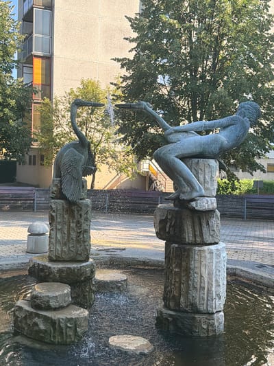 This is a vertical, eye-level, outdoor photograph of a tiered fountain featuring sculptures. The primary subjects are two bronze sculptures: one of a draped human figure, and another of a swan. The human figure, appearing to be female, is positioned on the upper tier of the fountain, seated and leaning forward with her arms extended. She appears to be pouring water from two vessels. The swan sculpture is on the lower tier, with its wings spread. Both sculptures are dark bronze in color and are mounted on stone bases with a carved, fluted design. Water is actively flowing from the upper sculpture, cascading down and creating a spray that is visible against the stone and in the air. The lower tier also shows water flowing.
The setting is a public space, likely a park or plaza, with a paved ground area and benches visible in the background. Tall green trees with dense foliage are prominent on the left side of the frame, partially obscuring a modern building with a light-colored facade and windows. The light suggests it is daytime, with sunlight casting shadows and highlighting the water. The overall mood is serene, with the movement of the water adding a dynamic element to the static sculptures. There is no visible text in the image. The location context provided is Tiszaszentmárton, Hungary.