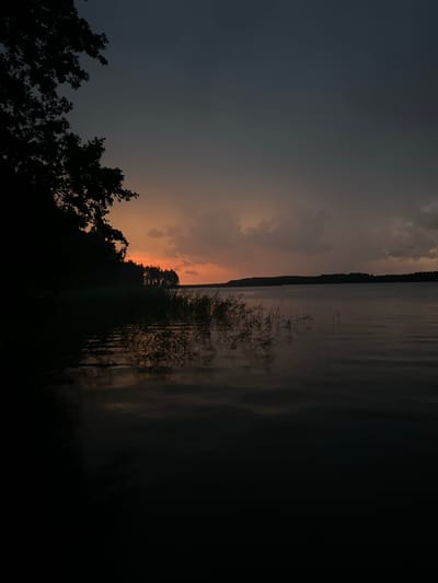 This is a vertical, low-angle, medium shot of a lake at dusk. The sky is a deep grey transitioning to a soft orange glow near the horizon. Dark clouds are visible in the upper right. Silhouetted trees and reeds line the left bank of the lake, their reflections appearing as dark shapes on the water's surface. The water itself is dark and appears calm, with subtle ripples. A dark, tree-lined landmass stretches across the horizon. The overall mood is serene and tranquil. This scene is located in Слабодкаўскі сельскі Савет, Belarus.