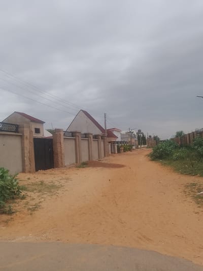 The image depicts a dirt road in the Gindi Nassarawa Kyarmi Galbi area of Nigeria. The road is unpaved and sandy, with tire tracks visible. On the left side of the road, there is a wall with decorative metal fencing and a large black gate. Behind the wall, houses with red roofs are visible. Further down the road, on both sides, are more houses and walls, some with solar-powered streetlights. Dense green foliage grows along the right side of the road. The sky is overcast with gray clouds. The scene appears to be a residential area.