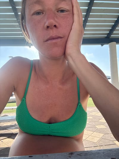 The image shows a close-up of a woman's face and upper body. She is wearing a green bikini top and has her hand on her cheek. She appears to be outdoors, possibly at a public park or beach, given the background which includes a tiled pathway and a grassy area. The lighting suggests it is daytime, with sunlight filtering through a slatted overhead structure. The woman's facial expression is neutral. The scene is set in Nahariya, Israel.