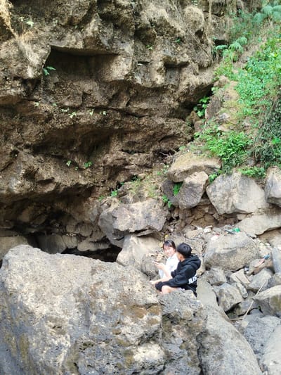 The image shows two individuals, a woman and a man, sitting on rocks in a rocky, natural environment. The woman is wearing a white top and the man is wearing a black hooded sweatshirt. They appear to be interacting, possibly talking or looking at something together. Behind them is a large rock formation, part of which forms a cave-like structure. Vegetation is visible on the right side of the rock formation. The environment is rocky and potentially rugged. There is evidence of trash amidst the rocks. The location is Đà Giang, Vietnam.