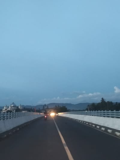 The image depicts a view from a moving vehicle on a bridge at dusk, likely in Cotcut, Indonesia. The bridge has white concrete railings with decorative elements. On the left side of the bridge, a white building with multiple domes, possibly a mosque or a large house, is visible in the distance. The road ahead has a white line marking its center. A motorcycle with its headlight on is approaching from the opposite direction. The sky is a pale blue with some scattered clouds, and mountains are faintly visible in the background. The overall atmosphere is serene and twilight-like.