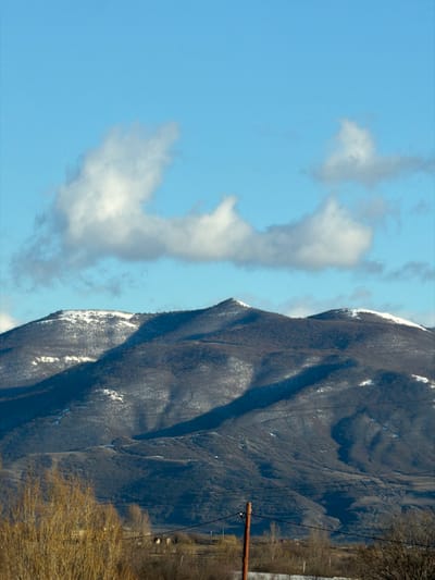 The image displays a mountainous landscape in Akhalsopeli, Georgia. The upper third of the frame is dominated by a clear, bright blue sky featuring scattered white cumulus clouds, with a prominent, elongated cloud formation stretching horizontally across the mid-sky. The central and lower-middle sections showcase dark, rugged mountains covered with sparse, brown vegetation, consistent with dormant deciduous trees. Patches of white snow are visible on the upper slopes and peaks, particularly on the highest peak positioned slightly left of center. Long, dark shadows are cast across the mountain faces, indicating a low sun angle. The foreground, at the bottom of the image, includes dry, leafless, brown vegetation on the left. A reddish-brown utility pole stands vertically slightly right of the center, carrying multiple dark power lines that extend horizontally across the lower part of the frame. Beyond the immediate foreground, an expanse of dry, yellowish-brown ground is visible, with several small, light-colored structures discernable in the distant midground. No individuals or specific actions are present in the scene.
