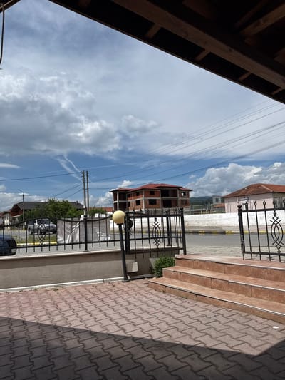 This is a daytime, outdoor image depicting a street scene in what appears to be a residential or developing area, located in Blatska, Bulgaria.
In the foreground, the edge of a dark wooden structure, possibly a roof or balcony overhang, is visible on the left, with multiple power lines stretching diagonally across the upper portion of the frame.
The midground features a multi-story unfinished building constructed from concrete, topped with a red tiled roof, indicating ongoing construction. To the right, a black wrought-iron fence with decorative motifs runs parallel to a paved sidewalk made of interlocking reddish-brown bricks. A street lamp with a spherical yellow-orange light fixture stands beside the fence. A set of stairs with metal edgings ascends from the sidewalk.
The background reveals another building with a red tiled roof, a paved road with distinct yellow markings, and several parked cars. Distant mountains are visible under a bright, partly cloudy sky. The sky is largely blue with significant white cumulus clouds and some faint, streaky contrails. Power poles and additional power lines are also present in the background.
There are no people visible in the scene. Notable details include the distinct shadows cast on the paved ground, indicating bright sunlight. A license plate "30 96 EH" is visible on one of the parked cars. A small white sign with partially illegible text, possibly "C 088...", is attached to the black fence.