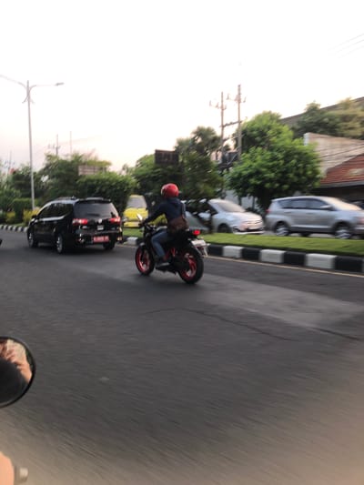 The image depicts a street scene in Klurak, Indonesia, captured during daytime with a slight motion blur. A motorcycle with a rider wearing a red helmet and jeans is moving from left to right across the frame. The motorcycle has red accents on its wheels. To the left of the motorcycle, a black SUV is also in motion, moving in the same direction. Further to the left, a street lamp is visible against a bright sky. In the background, there are trees and buildings, and other vehicles, including a yellow vehicle, are present on the road. The road is paved, and a grass median with black and white borders separates the lanes of traffic. The perspective suggests the photo was taken from another moving vehicle, possibly a motorcycle, as indicated by a partial view of a side mirror in the lower left corner reflecting a person's hand and arm.