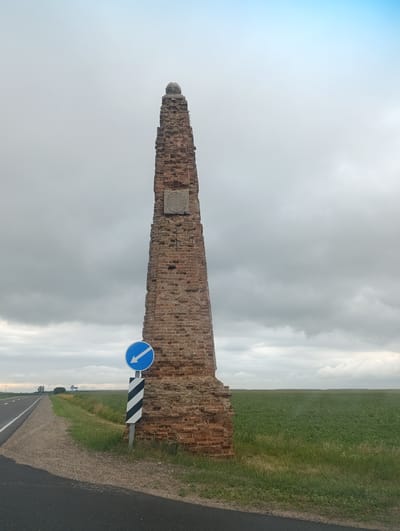 The image shows a tall, dilapidated brick obelisk situated beside a paved road in an open, rural landscape. The obelisk is weathered and crumbling, with visible damage to its structure. A circular blue road sign with a white arrow pointing left is attached to a pole in front of the obelisk, accompanied by a striped black and white marker. The road curves to the left, bordered by a grassy verge and a vast green field. In the distance, a small cluster of trees and a road sign with Cyrillic text are visible. The sky is overcast with a mix of grey clouds and patches of lighter sky, suggesting a possibly humid or cloudy day. The overall scene depicts a historical monument at the edge of agricultural land.