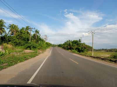 The image depicts a straight, asphalt road stretching into the distance under a blue sky with scattered clouds. The road is marked with broken yellow lines in the center and a solid white line on the left edge. Tropical vegetation, including palm trees, lines both sides of the road. Utility poles and overhead power lines are visible along the roadside. In the distance, a rice field landscape is also present. Based on the location in Payabeunyot, Indonesia, the surrounding area seems rural. A section of a vehicle is visible at the bottom of the image, indicating the photograph was taken from inside the car.