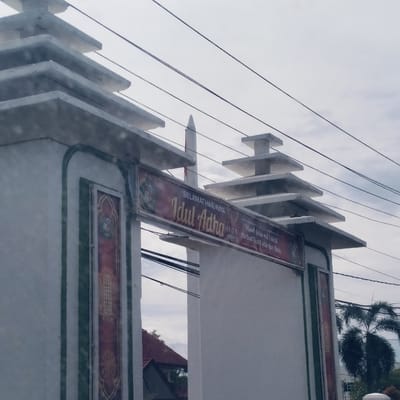 The image displays an ornate white archway or entrance gate, situated outdoors, likely in Lampeudeu Baroh, Indonesia. The archway features green trim along its edges and decorative panels with intricate patterns on its pillars. Each pillar is topped with multi-tiered, stepped architectural elements.

A red banner stretches across the top of the archway, bearing text in gold and white. The banner reads: "SELAMAT HARI RAYA Idul Adha," indicating the celebration of Eid al-Adha. Below "Idul Adha," the text "10/V H" and "2023 M" are visible, likely denoting the date of the holiday. To the right, additional greetings state "Minal 'Aidin wal Faizin" and "Mohon Maaf Lahir dan Batin," which are common Indonesian Eid greetings for seeking and granting forgiveness.

In the background, a tall, white pointed structure, resembling a minaret, rises against an overcast, cloudy sky. Several black electrical wires crisscross the upper portion of the frame. Through the archway on the left, a building with a reddish-brown tiled roof is partially visible. On the right, a large palm tree and other green foliage add to the natural elements of the scene. The image appears to be taken during the daytime under a soft, diffuse light from the cloudy sky. No people are present.