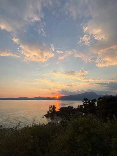 The image depicts a serene sunset over Lake Garda, Italy. The sun, a vibrant orange orb, is setting behind distant mountains, casting a warm glow across the water. The sky is a canvas of soft blues and pinks, with scattered clouds illuminated by the setting sun. In the foreground, lush green trees and foliage create a natural frame for the breathtaking view. A small beach area with scattered lounge chairs can be seen on the shore, suggesting a tranquil vacation spot. The overall atmosphere is one of peace and natural beauty.