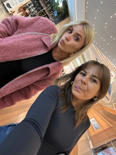 Two adult females are captured in a selfie-style photograph taken inside the Cap 3000 shopping center in Saint-Laurent-du-Var, France. The female in the foreground, positioned on the right, has medium-length brown hair with bangs, wears a dark long-sleeved top, blue denim jeans, and gold-colored hoop earrings. She is looking directly at the camera with a slight smile and her tongue extended. The second female, partially visible in the background on the left, has short blonde hair, wears a pink textured jacket over a black top, and gold-colored, abstract-shaped earrings. She is looking at the camera with her lips pursed into a pout and wide eyes. A silver-colored chain, likely from a purse, is visible over her left shoulder. The background reveals modern indoor architecture consistent with a shopping mall, including a light-colored slatted ceiling, curved structural elements, and reflective surfaces. A commercial sign bearing "Cap 3000" is partially visible in the upper left background, along with decorative lights from a distant establishment. The floor visible in the lower portion of the image is light-colored wood.