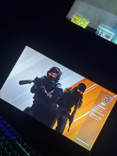The image displays a laptop screen illuminated in a dimly lit interior, located in Tsenovo, Bulgaria. The screen presents a video game or application interface featuring two stylized, armed figures. The left figure wears dark tactical gear, a helmet with an orange-tinted visor, and headphones, holding a suppressed firearm. The right figure is dressed in camouflage, a dark balaclava, and orange-tinted sunglasses, holding a rifle. The right side of the screen contains text in Bulgarian, a hexagonal emblem labeled "Mirage," and a progress bar at the bottom. A portion of a multi-colored backlit keyboard is visible in the lower-left foreground. In the upper-right background, two out-of-focus glass jars with swing-top lids, one yellow and one clear, are partially visible on a surface.