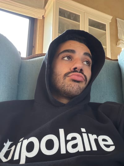An indoor, close-up photograph captures a single male individual, appearing to be in his 20s or 30s, seated in a light blue upholstered armchair. The individual has a medium-dark complexion, dark hair, brown eyes, and a moustache with beard stubble. He is wearing a black hooded sweatshirt with the hood pulled over his head. The word "bipolaire" is printed in white, stylized capital letters across the chest of the sweatshirt. The individual's head is slightly tilted upwards and to the right, with his eyes gazing off-frame in that direction, and his lips are pursed in a pout. The background shows an interior space featuring a bright window on the left, an adjacent light-colored wall, and a white wooden cabinet with glass doors displaying glassware, positioned above and behind the subject's head. The scene is located indoors in Mauléon-d'Armagnac, France.