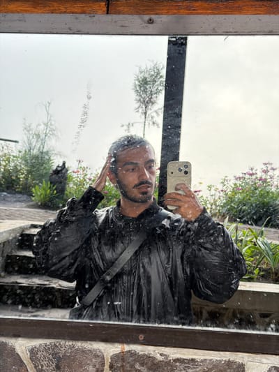 The image shows a mirror reflection of a young man taking a selfie. He is wearing a black, shiny, puffed-sleeve jacket and has a black strap across his chest. His hair appears wet, and he is touching his head with his right hand. He is holding a light-colored smartphone in his left hand, aiming it towards the mirror. The background reflected in the mirror shows lush greenery with small pink flowers, some trees, and a stone pathway with steps. The mirror itself is framed by dark wood and metal, and there are water droplets on the surface of the mirror, suggesting it might be raining or the environment is humid. The setting appears to be outdoors, possibly in a garden or a scenic outdoor area. Based on the provided location of Ngadas, Indonesia, this could be a reflection in a mirror at a hotel, restaurant, or tourist attraction in that area, known for its natural beauty and potentially humid climate.