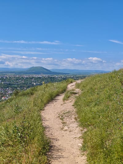 A dirt path winds up a grassy hillside under a bright blue sky with sparse white clouds. Below, a sprawling city is nestled in a valley, with a distinctive conical mountain visible in the distance. The city appears to be Goryachevodsky, Russia, given the landscape's resemblance to known geographical features of the area. The path is mostly clear, with some small rocks and sparse vegetation growing alongside it. The hillside is covered in green grass and wildflowers. The city itself is dense with buildings, surrounded by lush greenery. The overall scene depicts a sunny day with clear visibility.