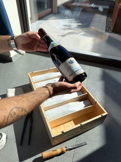 In Marche-lez-Écaussinnes, Belgium, an indoor scene depicts a person's two hands holding a dark glass bottle of "Paul Jaboulet Aîné La Chapelle Hermitage 1959" wine. The bottle features a dark red foil capsule and a main label with a blue and gold crest. The visible left forearm has a "wine" tattoo and a silver-colored watch. The visible right forearm is hairy, adorned with a thin gold-colored chain bracelet, and displays a partial tattoo. The bottle is held above an open, light-colored wooden wine crate labeled "LA CHAPELLE HERMITAGE," which contains four other bottles wrapped in white paper, laid horizontally in distinct compartments. Two compartments within the crate are empty. On the grey tiled floor, to the left of the crate, a pair of black-handled pliers or snips rests, and to the right, a wooden-handled chisel with a metal blade is present. The background consists of a large glass sliding door or window, reflecting an outdoor patio area with grey paving stones, dark planters, and landscaping elements, illuminated by bright sunlight which casts strong shadows on the indoor floor.