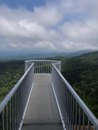 The image depicts a scenic overlook with a curved, white metal railing bordering a concrete pathway. Beyond the railing, a vast expanse of lush green hills and mountains stretches into the distance, partially obscured by a cloudy sky. The foreground is dominated by the overlook structure, creating a sense of depth and perspective. The scene suggests a natural landscape, likely a tourist attraction or a viewpoint. The location is specified as Nam Hải, Vietnam.