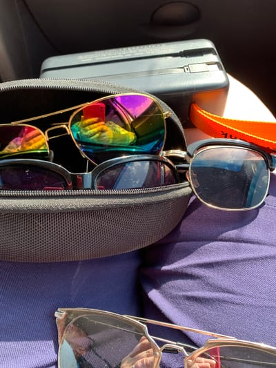 This is a close-up, top-down outdoor shot of a person's belongings, taken in the city of Азерскі сельскі Савет, Belarus. The primary subjects are several pairs of sunglasses, one pair with iridescent lenses and a gold frame, another with dark lenses and a black frame, and a third pair with clear lenses and a gold frame. The sunglasses are partially contained within a black zippered case, which is resting on a purple fabric surface, likely clothing. In the background, a gray electronic device with an orange strap is visible. The lighting suggests it is daytime, with bright sunlight casting shadows. The scene appears to be set within a vehicle, given the dark interior visible at the top of the frame. The reflection on the iridescent sunglasses shows a person holding a red smartphone.