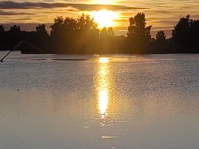 The image depicts a sunset over a body of water, likely a lake or river, in the vicinity of Усяжскі сельскі Савет, Belarus. Silhouettes of trees line the distant shore, with the sun partially obscured by clouds and casting a golden reflection on the water. A portion of a man-made structure, possibly part of a dock or a water sports facility, is visible on the left side of the frame. A small bird is seen flying in the lower left quadrant of the image. The overall scene conveys a serene, natural environment at dusk.
