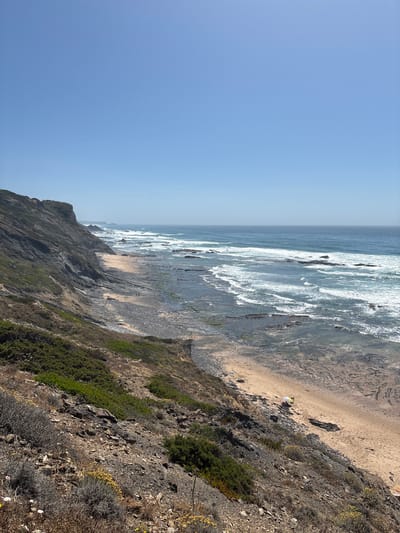 The image depicts a panoramic coastal scene on a clear, sunny day in Aljezur, Portugal. The sky occupies the upper portion, a gradient of light to medium blue, devoid of clouds. The horizon line, visible in the mid-ground, separates the sky from the deep blue Atlantic Ocean. The ocean exhibits multiple parallel lines of white-capped waves breaking towards the shore.

On the left, a prominent, steep coastal cliff dominates the mid-ground, composed of dark grey and brown stratified rock with visible erosion patterns. Sparse, low-lying green and dry scrub vegetation covers parts of the cliff face and the foreground slopes. The immediate foreground is a rocky, dry incline with various low-growing plants, including some with yellow and white flowers.

Below the cliff, a narrow sandy beach is exposed, interrupted by extensive dark, flat rock formations and tide pools. Two small, dark human figures are visible walking on a sandy section of the beach towards the upper left. Further down the beach, in the lower right quadrant, a small white and yellow parasol is open on the sand, next to what appears to be a person or an object. Another light-colored, possibly striped, beach umbrella is partially visible closer to the lower right edge. A prominent yellow and white paraglider canopy is laid out on a sandy patch of the beach in the lower mid-ground. The overall perspective is from an elevated vantage point looking down and along the coastline, extending into the distance where the cliffs and ocean gradually fade into a light haze.