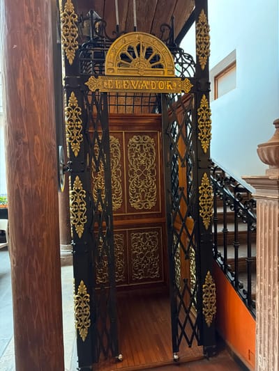 An ornate elevator gate, featuring gold filigree on black metal, spells out "ELEVADOR" vertically on its side. Intricate gold designs adorn the gate's bars, which form a lattice pattern. The elevator itself has dark wood panels with gold detailing. A half-circle wheel mechanism sits beside the lettering. Stairs are visible behind the elevator, leading upwards, adding to the historical feel.