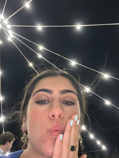 The image captures a close-up, low-angle selfie of a woman with dark hair and full lips, puckering her mouth and resting her right hand on her cheek. She is wearing a blue top, a large cross-shaped earring with colorful stones, and a gold ring with a black square stone on her right index finger. Her fingernails are painted a light blue. In the background, strings of small, bright lights are strung against a dark night sky, forming a radial pattern from the top left corner of the image. To the left and out of focus, a man in a light-colored shirt is partially visible. The scene appears to be outdoors at night, possibly in the city of Fasano, Italy, given the context.