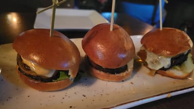The image shows three mini burgers, or sliders, arranged in a row on a speckled white rectangular plate. Each slider has a golden-brown bun, a dark patty, and appears to have some sort of sauce or cheese. The leftmost slider has lettuce and what looks like caramelized onions visible. The middle slider has a light-colored sauce. The rightmost slider has melted white cheese dripping down the sides. Each slider is secured with a wooden skewer. The plate is set on a dark, possibly wooden, surface. The background is blurred, with hints of ambient lighting and out-of-focus objects, suggesting an indoor setting like a restaurant. There are no people or animals visible in the image, and no specific location in Prévost, Canada, is identifiable from the visual information provided.