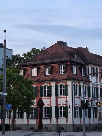 The image shows a corner building with a red-tiled mansard roof in Ansbach, Germany. The building has a white facade with red decorative bands and green shutters on the windows. There is a traffic light and a street sign visible on the left side of the image. A tree partially obscures the lower part of the building. A sculpture, resembling a stylized flame or seed, stands in front of the building. The sky is overcast with some clouds. The scene appears to be a street in a European city.