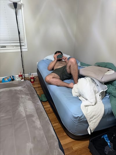 A medium shot presents the interior of a room in Franklin Township, United States of America. Dominating the center is an adult male, reclining on a large, blue inflatable mattress. He is fair-skinned, dark-haired and bearded, and is wearing dark-colored shorts. He is angled towards the viewer, and is looking at a dark-colored smartphone in his hands. The mattress is covered with a pale blanket.
Around the mattress, a variety of objects are visible. A black bag filled with dark clothes sits in the corner. Beside the mattress, several red cups and a water bottle are visible. Next to this cluster is a gray air mattress. White window blinds are visible on the left side of the frame.