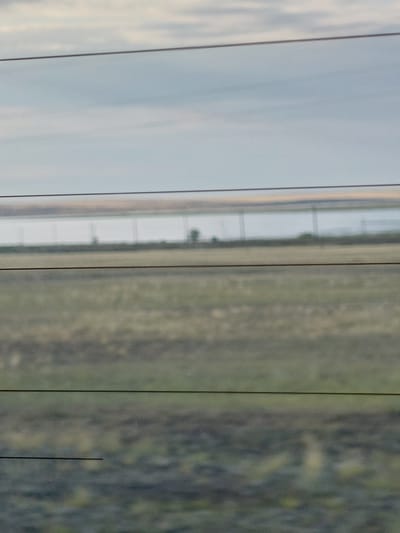 The image shows a landscape scene with a cloudy sky above a body of water and a field. There are thin, horizontal lines across the image, possibly indicating wires or a fence. The scene appears to be a rural or undeveloped area, with a uniform, low-lying horizon. There are no visible structures, people, or animals in the frame. The overall impression is one of open space and natural elements.