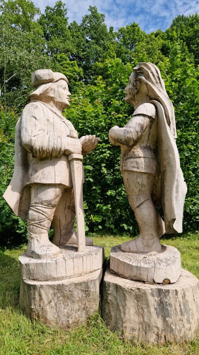 In Embūte, Latvia, two large, weathered wooden sculptures of figures stand side-by-side on tree stumps. The sculptures, appearing to be of men in historical attire, are positioned facing each other with their hands outstretched as if in conversation. The figure on the left wears a cap and cloak, holding a sword upright between its legs. The figure on the right also wears a cloak and appears to have long hair. Both figures exhibit carved details of clothing, including belts and boots. The backdrop consists of lush green foliage and trees under a partially cloudy blue sky. The foreground shows green grass and the rough texture of the wooden bases.