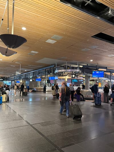 The image depicts a busy scene inside an airport terminal, likely Gardermoen, Norway, given the context. Several individuals are standing in line, each with their luggage, suggesting they are waiting for a flight or a baggage claim. The setting is modern, with a wooden-paneled wall on the left and a dark, tiled floor. Overhead, there are metal structures with various displays and lights.

The activity appears to be a typical airport waiting period. Some individuals are looking at their phones, while others are engaged in conversation or simply waiting patiently. The presence of multiple digital screens displaying flight information indicates an active travel hub. One screen shows "Oslo - 11:54" and another displays "Get 60kr off your first ride Uber". The overall atmosphere seems to be one of routine travel, with no apparent signs of distress or urgency. The lighting suggests it could be daytime, though the indoor setting makes it difficult to determine the exact time of day or weather conditions outside. The scene is well-lit, with a mix of overhead lighting and light emanating from the digital displays.