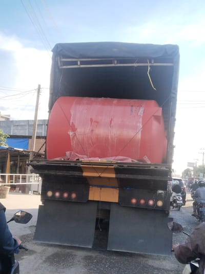 The image depicts a large, red water tank being transported on the back of a truck. The truck is covered with a black tarp, and the tank is partially visible underneath. The truck is on a street, with other vehicles and pedestrians in the background. The scene appears to be in a city, with buildings and utility poles visible. The sky is cloudy. The image is taken from a low angle, suggesting the photographer is on a motorcycle or in a low vehicle. The overall impression is one of daily life and commerce in a Southeast Asian city. Based on the context, this scene could be located in Blangpeuria, Indonesia, a region known for its agricultural and industrial activities.