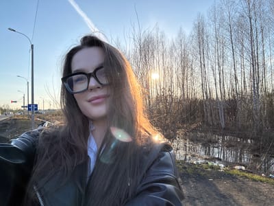 An outdoor selfie captures a young woman in what appears to be a semi-rural or roadside setting in Novoe Mart'yanovo, Russia. The woman, positioned on the right side of the frame, has long, dark brown hair partially obscuring her face. She wears black-rimmed glasses and a black leather jacket over a light-colored top. Her expression is neutral, and her lips are slightly parted. A strong sun flare emanates from behind her on the left, illuminating strands of her hair with a golden glow.

The background, under a clear blue sky with a visible contrail, is divided by a line of bare-branched trees, likely birches, whose reflections are visible in a calm body of water—possibly a ditch or pond—in front of them. The ground along the water's edge and what appears to be a path or roadside is composed of dirt and sparse green grass, noticeably littered with plastic bottles and other debris. To the right of the frame, utility poles supporting overhead lines and streetlights are visible. A blue traffic sign displaying two white arrows, one pointing straight and one pointing left, is attached to one of the poles. The bare trees suggest the image was taken during late autumn, winter, or early spring, with the low sun indicating either early morning or late afternoon.