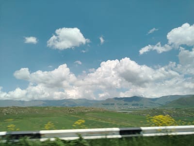 The image captures a scenic view of the landscape near Mirak, Armenia. The foreground features a white guardrail with blurred greenery in front, suggesting movement from a vehicle. Beyond the guardrail, a vast expanse of green fields stretches out, dotted with patches of yellow wildflowers. Rolling hills and mountains form the backdrop, with a small town visible nestled in a valley between them. The sky is a bright blue, filled with fluffy white cumulus clouds. The overall impression is one of a serene and picturesque rural setting under a clear sky.