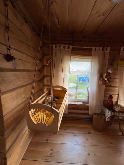 This image depicts a rustic, wooden room in Vybolovo, Russia, likely a bedroom or nursery. A wooden crib, suspended by ropes from the ceiling, hangs in the center of the room. The crib contains several stuffed animals and a yellow mattress. To the right of the crib is a window with white curtains, revealing a green outdoor scene with a blue vehicle visible in the distance. A small wooden table with a red pitcher and books stands to the right of the window. The walls and floor are constructed from natural wood, contributing to the room's cozy, traditional aesthetic. There are no people present in the image.