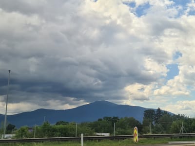 This image shows a mountain range with a large, dark blue mountain dominating the center. The sky above is filled with a mix of dark, heavy clouds and patches of bright, white clouds, with some blue sky visible. In the foreground, there is a road with a guardrail. To the left, a tall, grey pole stands vertically, likely a streetlight or traffic sign support. Along the edge of the road, there are green trees and bushes. A yellow and red striped traffic cone or marker is visible on the right side of the road. The scene appears to be a view from a roadside, possibly a highway, looking towards a mountain landscape on a cloudy day. This scene is consistent with the views found in and around Aichdorf, Austria, given its proximity to mountainous terrain.