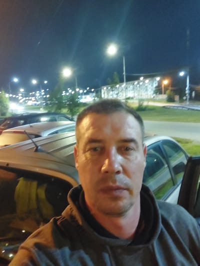A man is taking a selfie at night in a parking lot. He is wearing a gray hoodie. In the background, there are several cars parked, a building with many windows, streetlights, and trees. The scene appears to be in the city of Nakhabino, Russia.