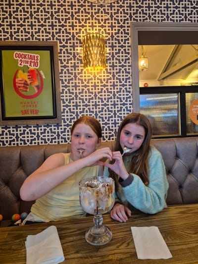 Here is a detailed description of the image:
**Scene:**
An interior shot, likely in a restaurant or pub setting.
**People:**
Two young girls are the focus. One is wearing a light green fuzzy sweater, and the other is in a yellow t-shirt. They are interacting with a large glass dessert dish. One of the girls is holding a spoon in her mouth and making a heart shape with her hands.
**Objects:**
- A large glass dessert dish, partially eaten, is on the table.
- Two white napkins are placed on the wooden table.
- A decorative lamp is affixed to the wall.
- A poster on the wall advertises "Cocktails 2 for £10" for Hungry Horse.
- Decorative wallpaper with a blue and white geometric pattern covers the wall.
- A tufted booth seat is visible behind the girls.
**Environment:**
The setting suggests an eatery of some kind with booth seating. The wall decor and poster point towards a casual dining or pub-like establishment.
**Inferences:**
- The image likely captures a moment of enjoyment or sharing of a dessert.
- The presence of the Hungry Horse poster suggests a location in the United Kingdom, as it is a British pub chain. While I cannot pinpoint the exact location in the Breckland District, the overall context makes that likely.
