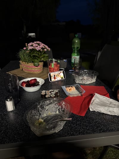 The image depicts a table setting outdoors at night. On the table are remnants of a meal including sushi, shrimp, salad in a plastic container, cherries in a white bowl, and watermelon in a container. A water bottle and a glass are present, as well as a salt shaker. A smartphone is displaying a photo of a meal, and a potted plant sits on the table. The location is likely in Hanau, Germany. Red napkins and white crumpled paper are also on the table. The table is dark gray with a speckled pattern. A chair and grass are partially visible at the edge of the table. The overall scene suggests a casual outdoor dining setting.