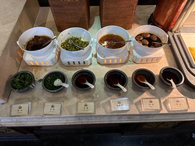 An indoor culinary display counter is depicted in Tongxiang, China, presenting various Chinese food items and condiments. The counter surface is light-colored and speckled.

In the upper tier, four large white ceramic bowls rest on individual illuminated white warming stands. From left to right, these contain: dark stewed meat in a thick brown sauce with a metal serving spoon; finely chopped green vegetables or herbs with a metal serving spoon; a reddish-brown stew or sauce with some solids and a metal serving spoon; and dark braised eggs in a dark liquid with a metal serving spoon.

In the lower tier, five smaller, dark gray stone-like bowls are arranged. From left to right, these contain: fresh chopped green coriander with a small metal scoop; chopped green scallions with a white ceramic spoon; a dark reddish-brown liquid (labeled as "Rice Vinegar") with a white ceramic spoon; a lighter brown liquid (labeled as "Sesame Oil") with a white ceramic spoon; and a dark brown liquid (labeled as "Soy Sauce") with a white ceramic spoon. Each small bowl is accompanied by a clear acrylic label displaying its Chinese name and English translation.

In the background, a brown wicker-patterned element is visible. To the far right, a portion of a metal structure and a white tray holding a yellow food item indicate a larger buffet setup. No individuals, actions, or direct interactions are observed in the scene.